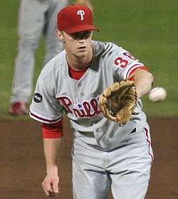 A man in a gray baseball uniform with red trim and "Phillies" across the chest in red script and wearing a red baseball cap with a white "P" on the front reaches for a baseball with his baseball glove