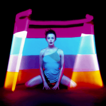 A body image of a woman (Kylie Minogue) inside of a cut cone with multi-colored lights. From top: Purple, blue, mid-purple/blue, oranges and red. The woman is wearing a small blue mini dress with detail on the top left, whilst the floor has a reflection of the cone.