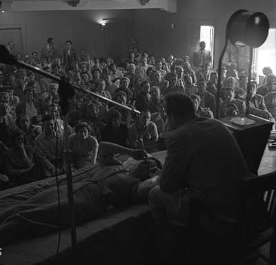 A mostly seated crowd watches as Hubbard, seated on a chair, speaks to a woman lying prone in front of him.
