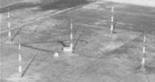 Aerial view of five tall antenna towers standing on flat terrain; four are arranged in a square, and the fifth one is at the center