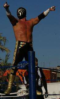 Wrestler La Sombra standing on the second rope of a wrestling ropes, pointing to the fans during an outdoor event.