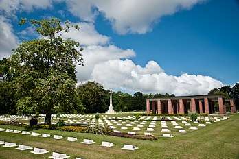 Colour photo of a cemetery comprising rows of small stone grave markers. A cross-shaped monument and a red brick structure are visible in the background