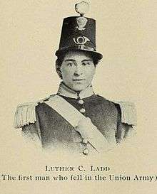 A sepia toned portrait photograph depicting the head and shoulders of a young man in an elaborate militia uniform. He wears a tall dress uniform hat.