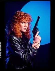 An image of a woman with bright red, curly hair. She is holding a gun and looking toward the camera.