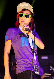 A woman in a pink t-shirt and black skinny jeans with sun glasses and a baseball cap with a "Bobbed" haircuit singing into a microphone