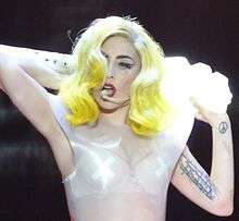 Gaga performing with her hands raised behind her neck. On the left biceps, a line of tattoos is visible.