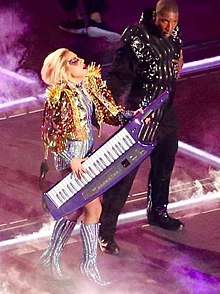 Lady Gaga playing a keytar while wearing a gold jacket. Richy Jackson can be seen to her left.