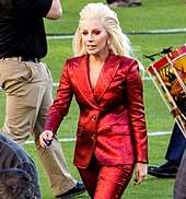 Lady Gaga in a red pant-suit walking on a field.