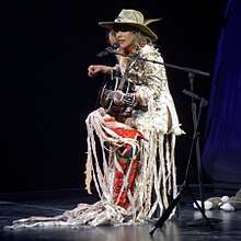 Gaga wearing a white dress, red boots and a hat, playing guitar onstage