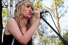 A woman wearing a black singlet and pink hairband, singing into a microphone.