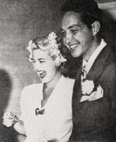Man and woman smiling; the woman has flower arrangements in on each side of her head