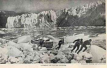  Six men pulling a boat onto an icy shore, with a line of ice cliffs in the background