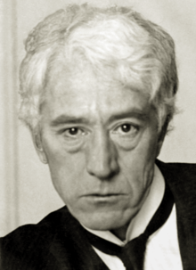A white-haired man in his fifties, wearing a black jacket and tie and white shirt.