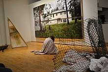 Lara is covered by a dirtied white sheet sitting on the floor. To her right is half of a canoe, and her left a rolled up snow fence. Behind her is a large image of Pelican Lake residential school.