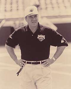 Candid sepia-toned photograph of Pasquale wearing a black polo-style shirt and white baseball cap both bearing the logo of the Jacksonville Jaguars and standing on a football field with arms akimbo