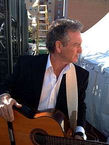 A grey-haired man wearing a black jacket and white shirt, holding a guitar