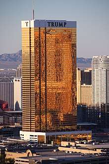 A tall rectangular-shaped tower in Las Vegas with exterior windows reflecting a golden hue. It is a sunny day and the building is higher than many of the surrounding buildings, also towers. There are mountains in the background.