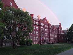 A long, five-story brick building stands in the evening light with a rainbow overhead