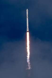 A white Falcon 9 rocket cuts through the blue sky, with its nine engines producing a bright yellow flame