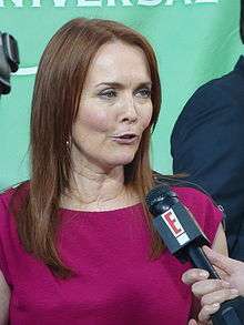A lady with long brown hair and a red shirt in front of a E! microphone, and she is talking.