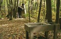 A metal sink with running water supported by wooden beams in the foreground, a wooden open shelter with a stone chimney amidst the trees in the background