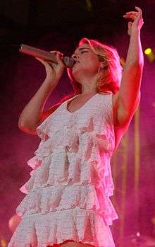 A young blonde-haired woman wearing a white dress, singing into a microphone