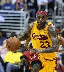 Black man dribbling a basketball with a gold and maroon basketball jersey that says Cavaliers in maroon cursive lettering across the chest. He is wearing a headband with an NBA logo.