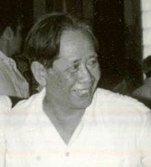 A smiling man looking to the right, wearing a collarless white shirt