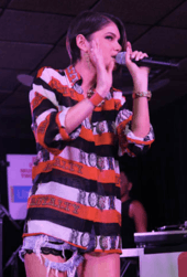 An image of a young woman performing on stage. She is wearing a red, white, and black striped shirt and denim shorts.