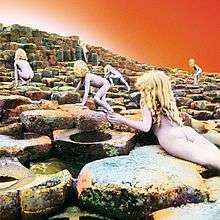 Six nude children with long blonde hair scramble up a stairstep series of basalt rocks ascending away from the viewer, with an orange-white sky above