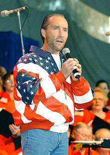 A middle-aged man wearing a jacket with an American flag design and blue jeans, singing into a microphone