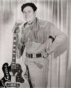A dark-haired man wearing a neckerchief, a shirt with fringes dangling from the sleeves, and pants with a guitar pictured on them, smiling broadly while leaning on a guitar