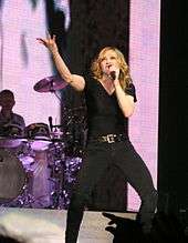 A female blond performer singing to a microphone while lifting her left arm.