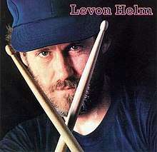 Head shot of Levon Helm, wearing a blue cap and holding two drumsticks in front of his face