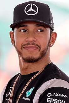 A man in his early thirties wearing a black baseball cap and t-shirt. He has a gold neck chain across his body.