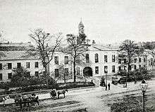 A black and white photograph, showing a stately-looking two storey building with white walls, extending out of shot to the left and right, with an arched cart entrance at the centre. A modest clocktower rises above the entrance, and the building is surrounded by neat shrubbery and iron railings. A wide street crosses left-right outside of the fence, with a handful of horse-drawn carts and pedestrians in 19th century clothing.