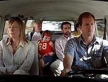 A film screenshot shows the family all seated in the Volkswagen Microbus as it is driven on a highway. The angle is from the windshield looking into the vehicle so that the majority of the interior can be seen. The mother is sitting in the front passenger seat with a dull expression on her face. The father is driving the vehicle with a similar expression. In the middle row, the daughter is looking down, listening to music from a CD player. Her uncle is seated on the right, looking to his left. In the back row, the grandfather is looking towards his grandson (whose face is slightly obscured by his father's head due to the angle). In the background, other vehicles can be seen driving on the highway.