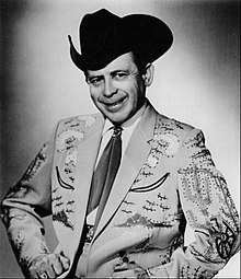 A smiling man wearing a dark cowboy hat and a light-coloured jacket with an elaborate pattern on it