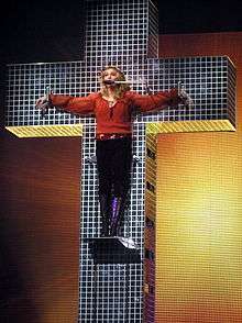 An image showing a blue crystal cross and a blond woman standing on a platform on the cross. The woman is wearing a red shirt and dark brown pants. Her hands are spread apart along the cross's breadth to symbolize as if she has been crucified. Behind the cross, a backdrop is centrally illuminated