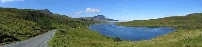 A blue body of water sits beneath a blue sky surrounded by green moorland. A road to the left travels along the lake side leading towards a small patch of mist and some low hills in the distance.