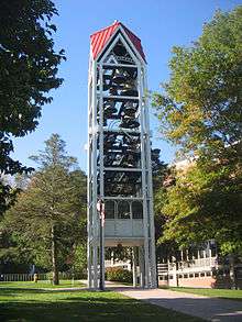 A three-dimensional vertical structure perhaps 70 feet (21&nbsp;m) tall and 10 feet (3.0&nbsp;m) wide by 10 feet (3.0&nbsp;m) long is filled with tiers of large metal bells. The structure, standing in front of a large institutional building in a park-like setting, is supported by several metal legs. A sidewalk passes through the structure, which is open at the bottom to a height of perhaps 10 feet (3.0&nbsp;m).