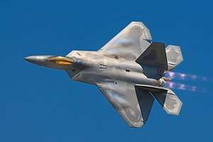 Dorsal view of F-22 in steep bank, ailerons clearly banking wings, modern camouflage, bright gold canopy, jet thrust possibly purple with afterburner and cloud-effect from leading edges of wings all suggesting high speed