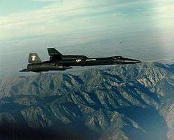 Sideview of black jet aircraft overflying mountain towards right of photo.
