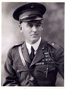 A black and white image of Logan Feland, a white male in his Marine Corps Service A uniform. He is wearing a hat several ribbons and a Fourragère. He also has a leather strap from his right shoulder to his left waist.