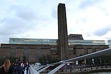 A large oblong brick building with square chimney stack in centre of front face. It stands on the far side of the River Thames, with a curving white foot bridge on the left.