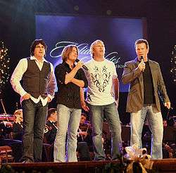 Four middle-aged men standing on a stage in front of an orchestra.  Two are holding microphones.