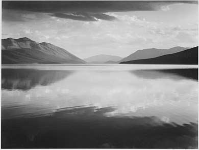 A black-and-white photograph shows a large, still lake extending horizontally off the frame and halfway up vertically, reflecting the rest of the scene. In the distance, a mountain range can be seen, with a gap in the center and one faint smaller mountain in between. The sky is cloudy and large dark clouds rest at the very top of the frame.
