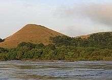 A view of a wide river in the foreground, with a single dirt mountain surrounded by trees in the background.