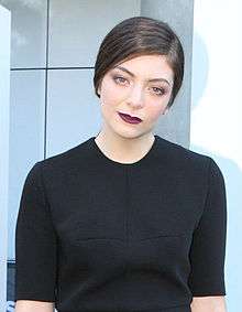 Lorde is wearing a black dress as she faces the camera.
