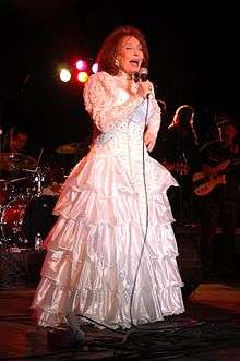 An older woman with long brown hair wearing a long white dress, singing into a microphone with musicians behind her